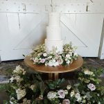 Stunning white three tier wedding cake with pearl decoration to all layers surrounded by a floral wreath on the table