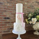 Tall three tier wedding cake with a ruffle iced layer, a layer decorated with the couple's initials, and a layer decorated with pearls and a pink ribbon draped down the cake