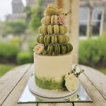 White iced wedding cake with hand painted pastel green decor and floral decoration topped with a stack of Macarons in shades of green