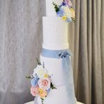 Tall five tier white wedding cake with pastel blue and pink floral decor and a baby blue ribbon to the central tier